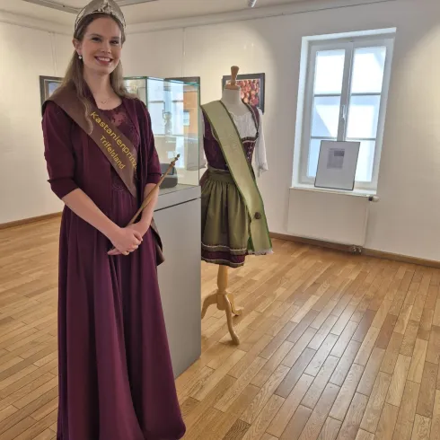 Eine Frau in einem burgunderfarbenen Kleid steht lächelnd in einem Ausstellungsraum. Im Hintergrund ist ein Kleid auf einer Schneiderpuppe zu sehen.