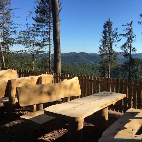 Ein Holzsitzecke mit Tischen und Bänken in der Natur. Im Hintergrund sind Bäume und eine hügelige Landschaft zu sehen.