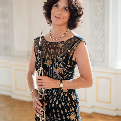 Eine elegante Frau steht in einem geschmackvoll dekorierten Raum. Sie hält ein Oboe und trägt ein schönes, verziertes Kleid.