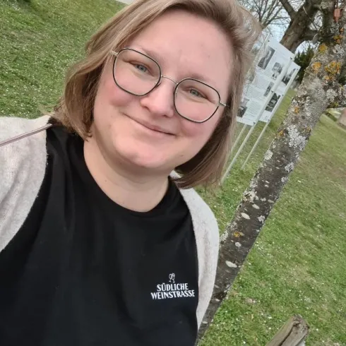 Eine Person mit Brille lächelt vor einem Grünen Hintergrund. Im Hintergrund sind Informationsschilder zu sehen.