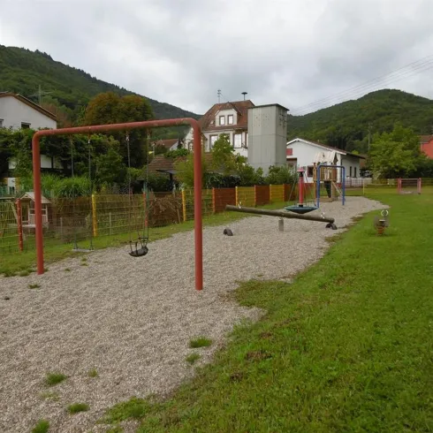 Ein Spielplatz mit Schaukeln und einer Wippe auf kiesigem Boden. Im Hintergrund sieht man Häuser und grüne Hügel.