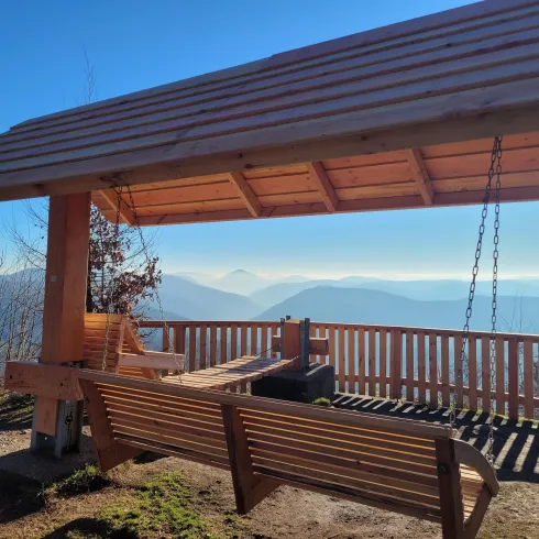 Eine charmante Aussichtsplattform mit Holzschaukeln und Dach. Im Hintergrund erstrecken sich sanfte Berge unter einem klaren Himmel.