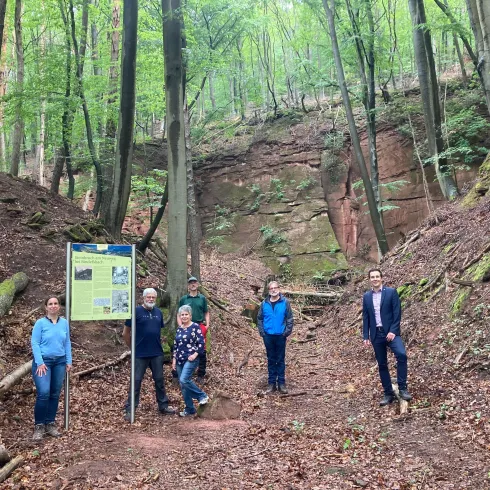 Die Gruppe von Menschen steht im Wald an einem Wegweiser. Der Boden ist mit Laub bedeckt und die Umgebung ist grün und bewaldet.