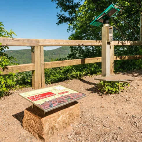 Eine Aussichtsplattform mit einem Panoramaschild vor einer grünen Landschaft. Der Himmel ist klar und die Umgebung ist von Bäumen umgeben.