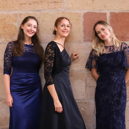 Drei Frauen in eleganten, dunkelblauen Kleidern posieren vor einer Steinmauer. Sie lächeln und haben unterschiedliche Frisuren.