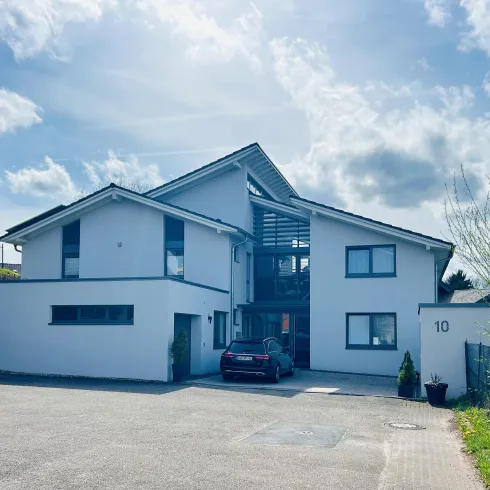 Ein modernes, weißes Haus mit großen Fenstern und einem begrünten Vorgarten. Der Parkplatz vor dem Haus ist offen und ein Auto steht dort.