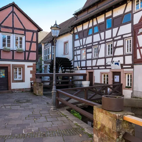 Ein malerischer Platz mit Fachwerkhäusern in hübschen Farben. Im Hintergrund ist ein kleiner Wasserlauf und eine Straße zu sehen.