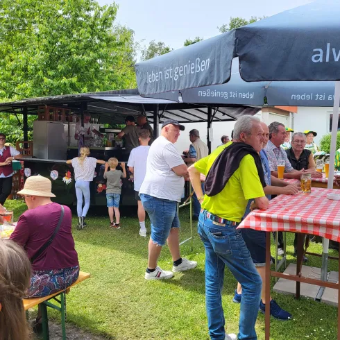 Eine lebendige Gartenparty mit vielen Menschen, die an Tischen sitzen und trinken. Es gibt einen Foodstand im Hintergrund und viel Grün um die Veranstaltung herum.