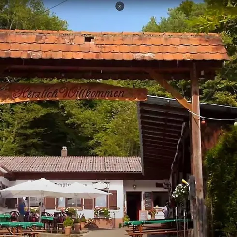 Ein einladendes Holztor mit der Aufschrift "Herzlich Willkommen". Im Hintergrund ist ein Gasthaus umgeben von Bäumen und Sitzplätzen im Freien zu sehen.