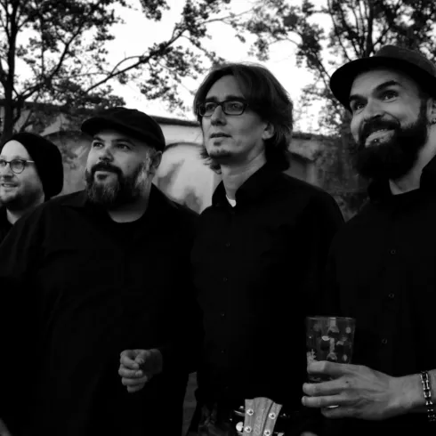 Eine Musikgruppe steht im Freien unter Bäumen. Alle Mitglieder tragen schwarze Kleidung und lächeln in die Kamera.
