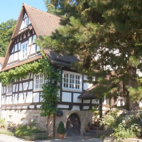 Ein traditionelles Fachwerkhaus mit grünen Pflanzen und einem einladenden Eingang. Die Umgebung ist von Bäumen und einer ruhigen Straße geprägt.