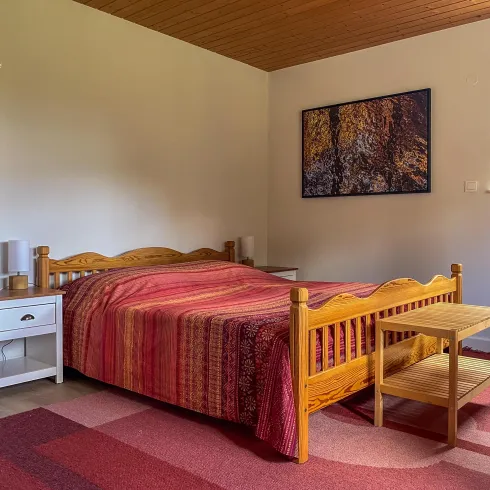 Ein gemütliches Schlafzimmer mit einem Holzbett und einer bunten Bettdecke. Die Wände sind hell und es gibt ein Fenster mit Blick nach draußen.