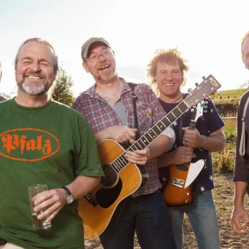 Eine Band von fünf Männern steht lächelnd im Freien. Sie halten Musikinstrumente und wirken entspannt und freundlich.