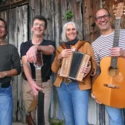 Eine Gruppe von vier Musikern steht lächelnd vor einer alten Holzwand. Sie halten verschiedene Musikinstrumente, darunter eine Flöte, eine Gitarre und ein Akkordeon.