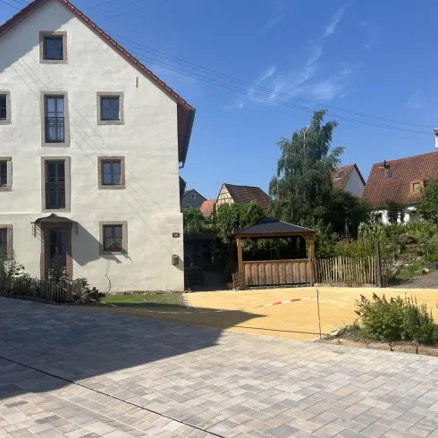Ein großes, helles Haus steht in einer ruhigen Umgebung. Im Vordergrund ist ein gepflasterter Weg zu sehen, neben einem Garten mit einer Pergola und viel Grün.