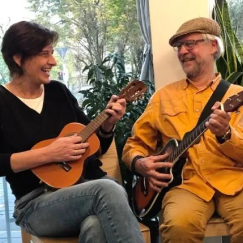 Zwei Personen sitzen zusammen und spielen Gitarre und Ukulele. Beide lächeln und haben Spaß beim Musizieren.
