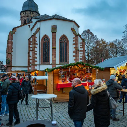 Ein Weihnachtsmarkt mit festlich dekorierten Ständen und vielen Besuchern. Im Hintergrund ist eine Kirche zu sehen, die die winterliche Atmosphäre unterstreicht.