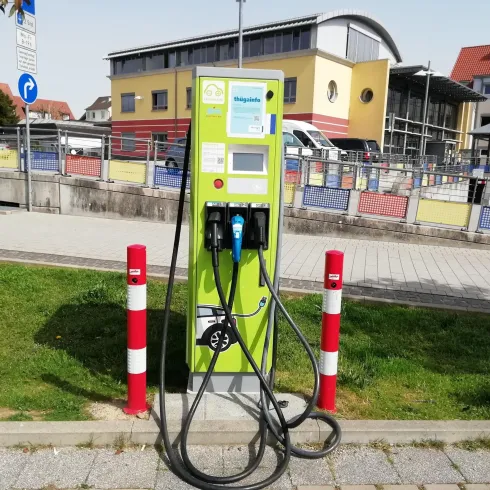 Eine Elektroladestation steht auf einem Gehweg. Um die Station sind grüne Rasenflächen und einige Gebäude zu sehen.