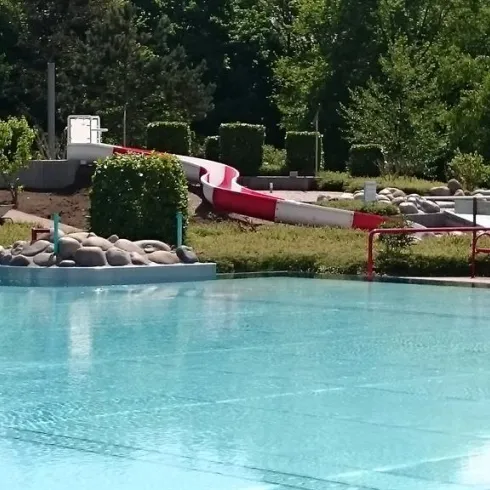 Ein Schwimmbad mit klarem, blauen Wasser und einer Rutsche im Hintergrund. Umgeben von grünen Pflanzen und Büschen bietet es einen einladenden Ort zum Entspannen.