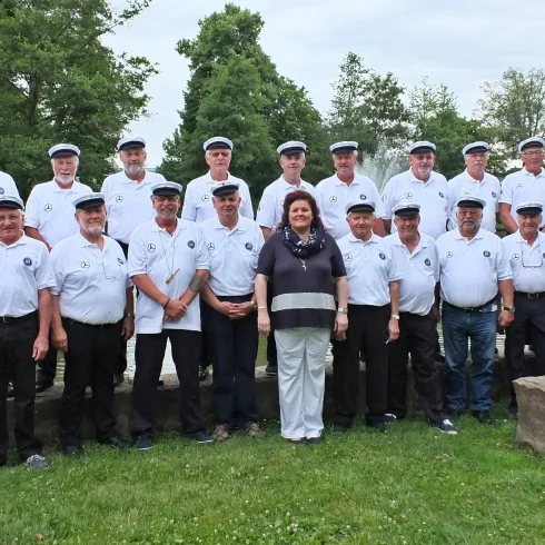 Eine Gruppe von Männern in weißen Hemden und Hüten steht auf einer Wiese. In der Mitte steht eine Frau in einem schwarzen Oberteil.