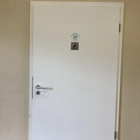 Eine weiße Tür mit einem Schild und einer Kamera darüber. Die Wand ist in einem schlichten Farbton gehalten.