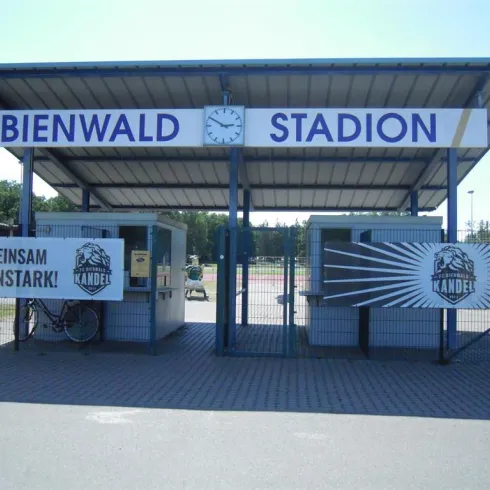Ein Stadion mit dem Namen "Bienwald" ist zu sehen. Vor dem Eingang hängen Schilder mit Aufforderungen zur Unterstützung der Mannschaft.