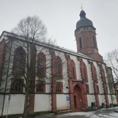 Ein historisches Gebäude mit einem hohen Turm. Die Fassade ist in rot und beige gehalten, umgeben von Bäumen und einer ruhigen Straßenszene.