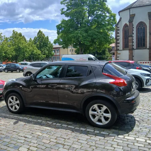 Ein schwarzes Auto steht auf einem Kopfsteinpflasterparkplatz. Im Hintergrund sind Bäume und einige Gebäude sichtbar.
