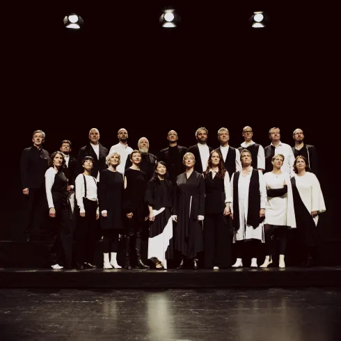 Eine Gruppe von Sängern steht auf einer Bühne, alle in schwarz und weiß gekleidet. Der Hintergrund ist dunkel, und es gibt ein paar Scheinwerfer, die die Gruppe beleuchten.