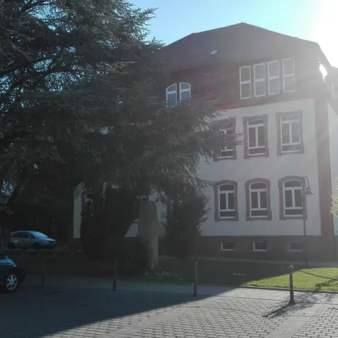 Ein Gebäude mit mehreren Fenstern, umgeben von Bäumen. Im Vordergrund steht ein parkiertes Auto.