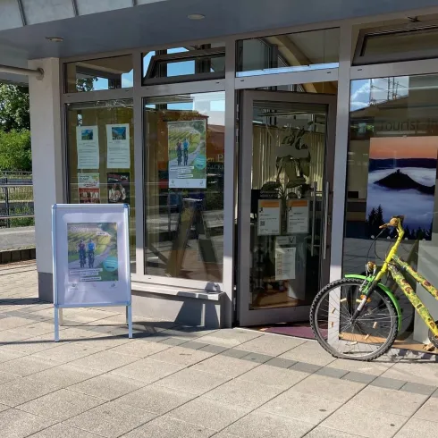 Ein Geschäft mit großen Fenstern und einem Eingang. Vor der Tür steht ein gelbes Fahrrad mit einem Korb und einem Werbeschild.