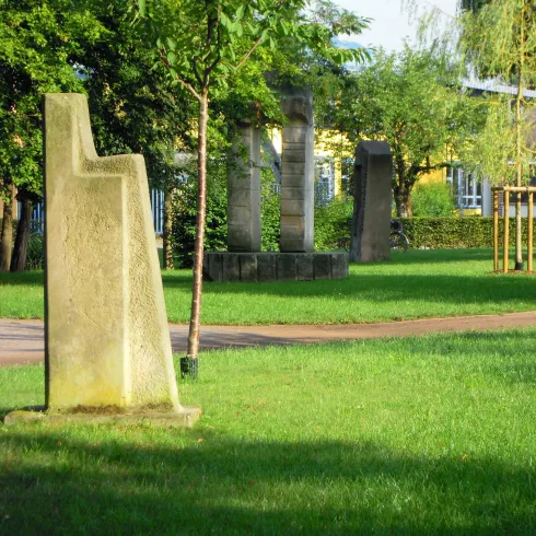 Ein ruhiger Park mit grüner Wiese und Bäumen. Im Vordergrund steht eine markante Steinskulptur.