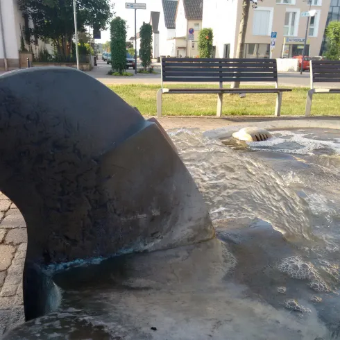Ein Teil eines Brunnens mit einer skulpturalen Welle und sprudelndem Wasser. Im Hintergrund sind Bänke und grüne Rasenflächen zu sehen.
