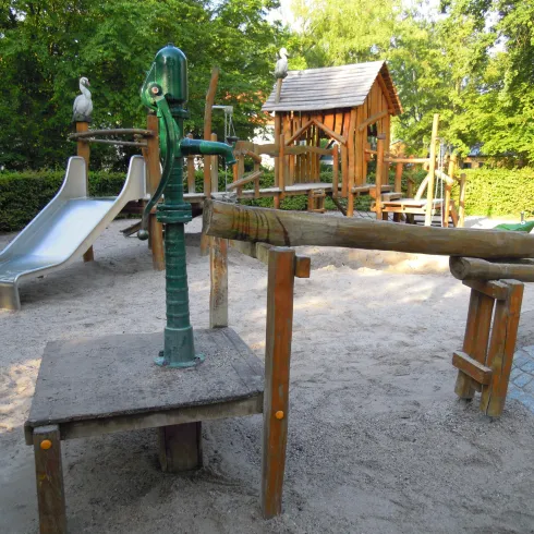 Ein Spielplatz mit einer Rutsche, einer Pumpe und einem Holzhaus. Der Bereich ist mit Sand und Bäumen umgeben.