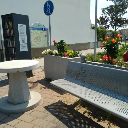Eine moderne Sitzgelegenheit mit einem Tisch und einer Bank steht in einem blühenden Garten. Im Hintergrund sind bunte Blumen und ein Verkehrsschild zu sehen.
