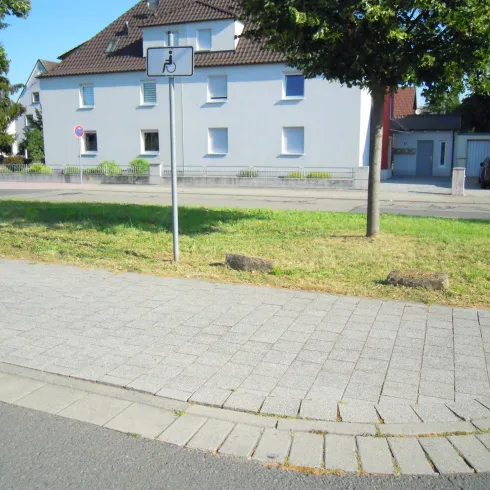 Eine ruhige Straßenszene mit einem Basketballkorb und einem grauen Gebäude im Hintergrund. Im Vordergrund gibt es einen gepflasterten Gehweg und einen kleinen Grünbereich.