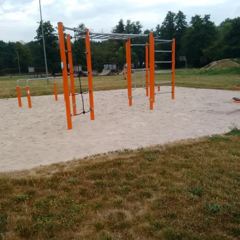 Ein Spielplatz mit orangefarbenen Fitnessgeräten auf einem Sandbereich. Im Hintergrund sind Bäume und eine Grünfläche zu sehen.