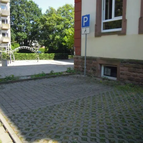 Ein ruhiger Parkplatz neben einem Spielplatz. Im Hintergrund sind Bäume und ein mehrstöckiges Wohnhaus zu sehen.