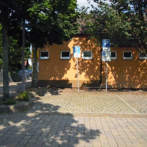 Ein Parkplatz mit verschiedenen Verkehrsschildern und einer orangefarbenen Wand im Hintergrund. Die Umgebung ist grün mit Bäumen und auf dem Boden sind Pflastersteine.