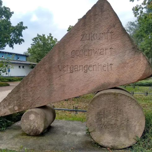 Ein monumentales Steinrelief mit der Aufschrift "Zukunft - gegenwart - vergangenheit". Es steht im Freien, umgeben von Bäumen und Grünflächen.