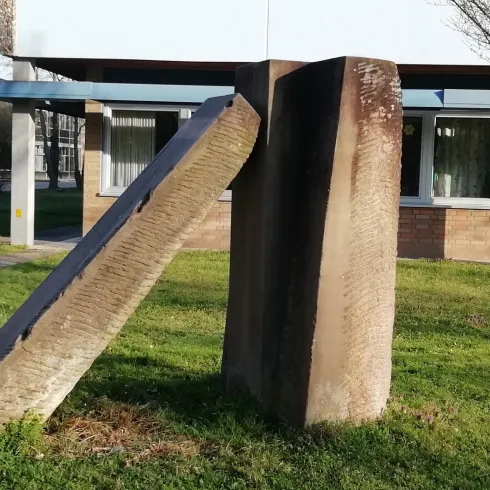 Eine abstrakte Skulptur aus Beton steht auf einer grünen Wiese. Sie besteht aus zwei schrägen, aufeinander gestellten Blöcken.