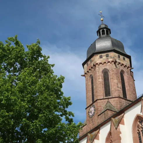 Ein hoher Kirchturm mit einer charakteristischen Kuppel. Daneben steht ein großer, grüner Baum unter einem blauen Himmel.