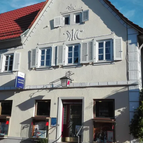 Ein historisches Gebäude mit hellgrauer Fassade und roten Fensterläden. Die Fenster sind freundlich dekoriert und das Dach ist rot.