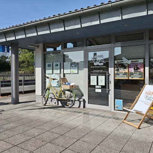 Ein modernes Informationszentrum mit großen Fenstern und einem Fahrrad vor der Tür. Auf dem Bürgersteig steht ein aufklappbarer Gästeständer mit Informationen.