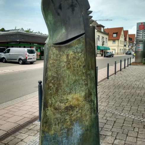 Eine abstrakte Skulptur aus Metaller mit einer grünlichen Patina. Sie steht auf einem Gehweg in einer städtischen Umgebung.