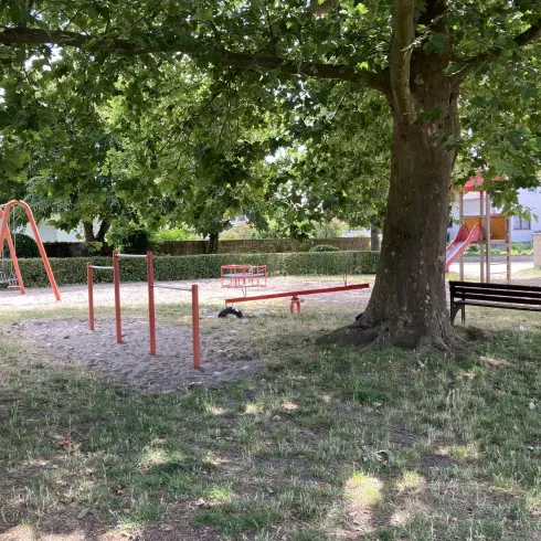 Ein Spielplatz mit einer Schaukel, einem Klettergerüst und einer Bank steht unter einem großen Baum. Die Umgebung ist grün und einladend.