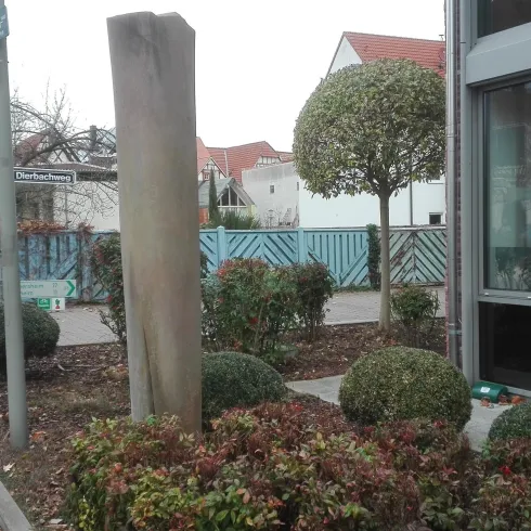 Eine straßenseitige Ansicht mit einem großen Stein und gepflegten Büschen. Im Hintergrund ist ein modernes Gebäude zu sehen.