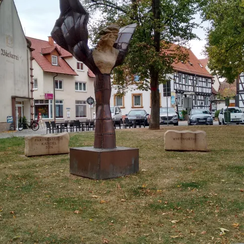 Eine Skulptur steht in einem Park, umgeben von Bäumen und Gebäuden. Der Kunstwerk zeigt eine Figur, die einen Vogel hält.