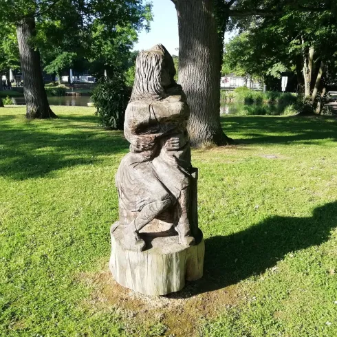 Eine geschnitzte Holzskulptur eines sitzenden Menschen auf einem Baumstamm. Umgeben von frischem Grün und Bäumen im Hintergrund.