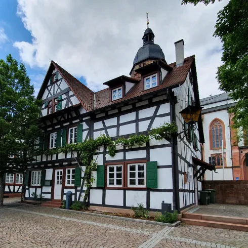 Ein gemütliches Fachwerkhaus mit einem hohen Dach und grünem Fensterladen. Im Hintergrund sind Bäume und ein Teil einer Kirche sichtbar.